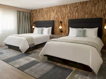 A modern hotel room with two queen beds, wooden feature wall, bedside lamps, light bedding, and a patterned rug.