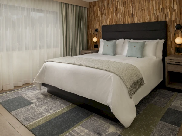 A stylish hotel bedroom with a large bed, soft white linens, mint throw pillows, and warm pendant lights beside nightstands.
