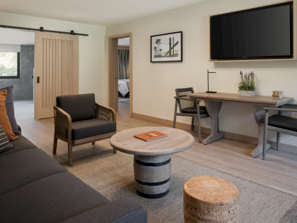 A modern living room with a dark sofa, two armchairs, round coffee table, textured rug, wall-mounted TV, and a desk with a chair, plus a small round cork stool.
