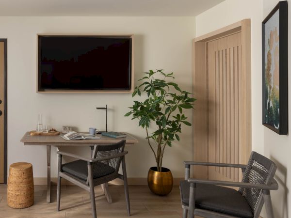 A modern office nook with a wooden desk, chairs, books and coffee cups, a potted tree, framed art, and a door.