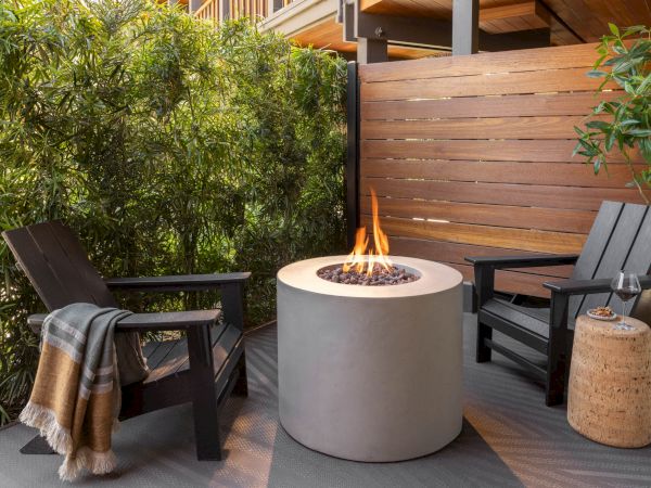 Outdoor patio scene with a lit fire pit, dark chairs, and a wood privacy wall beside lush bamboo plants.