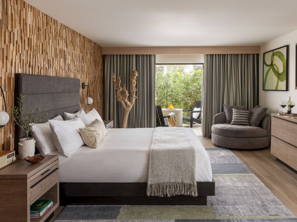 A modern hotel-style bedroom with a large bed, gray sofa, wood wall, curtains, and outdoor greenery view through the window.