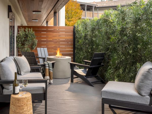 An outdoor patio with cushioned chairs, a tabletop wine bottle, and a flaming fire pit beside lush greenery.