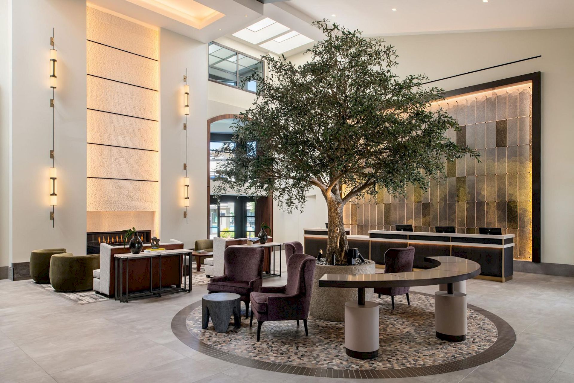 A modern hotel lobby with a central tree, circular seating around a polished tree feature, contemporary art wall, soft lighting, and neutral tones.