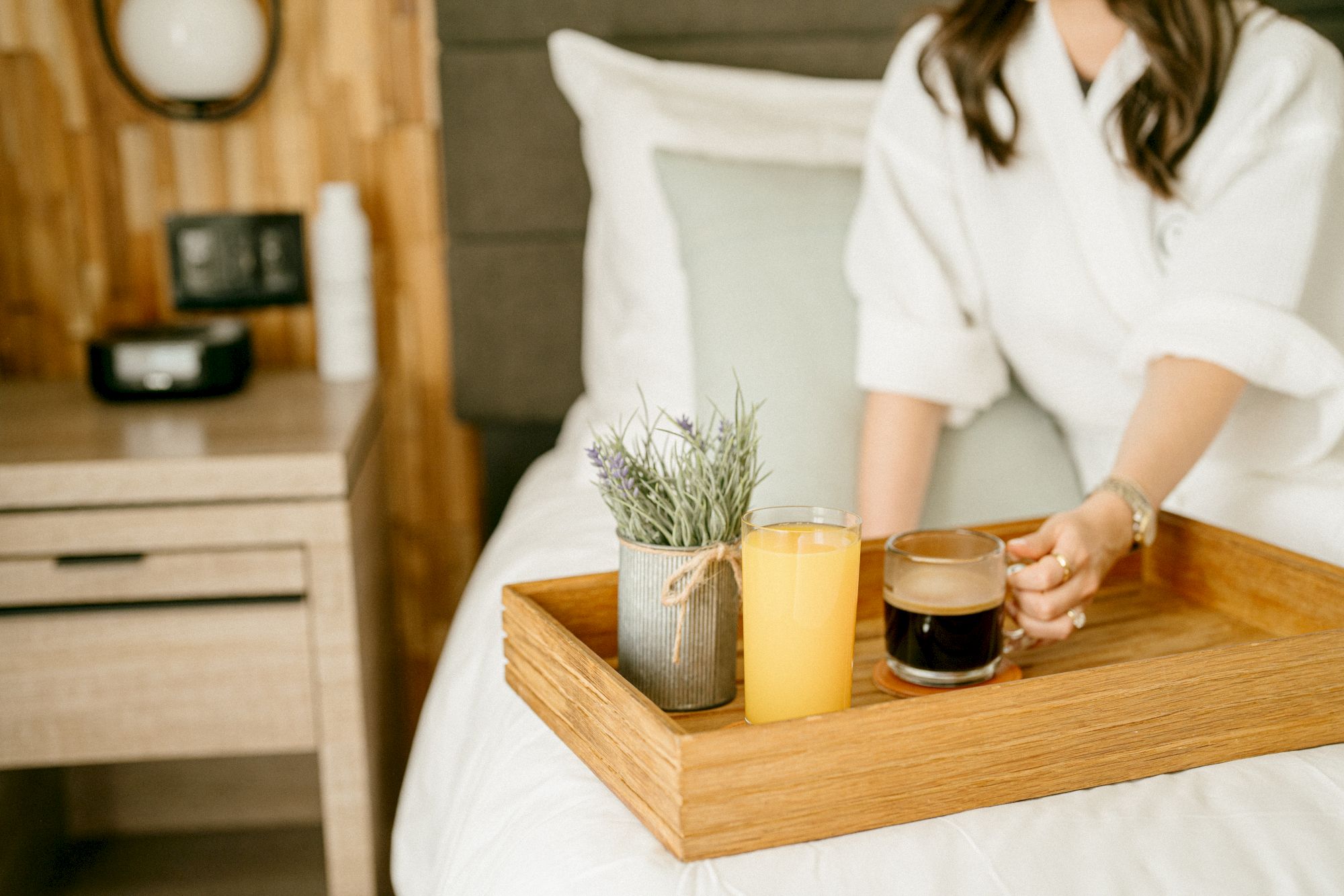 Person on a bed wearing a robe, holding a wooden tray with a glass of orange juice, a mug of coffee, and a small plant; breakfast in bed.