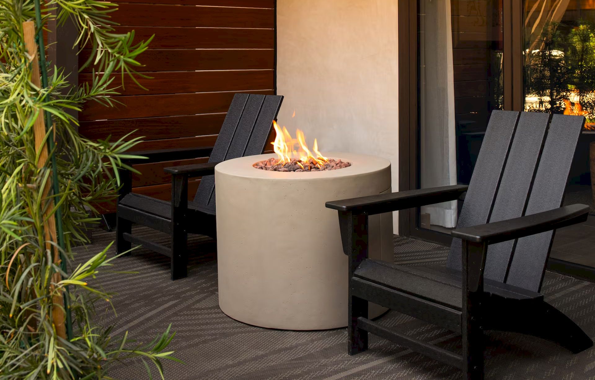 A small patio with two black wooden chairs around a round fire pit, flames crackling, and plants along the edge.