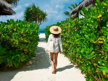 A woman strolling through a sunny tropical path, wearing a wide-brim hat and white cover-up, heading toward a blue ocean waves.