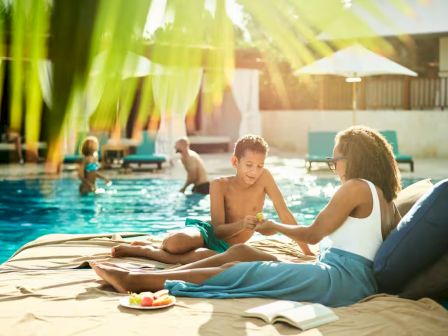 People relaxing by a sunny poolside. A woman and child sit on lounge chairs, chatting, with towels, books, and snacks nearby. Other swimmers in the background.