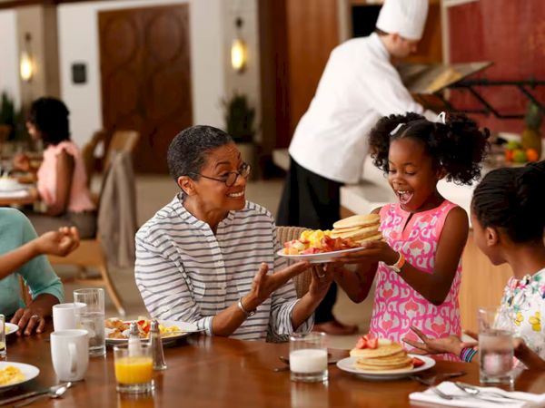 Friends share a cheerful breakfast in a busy restaurant, passing a plate of pancakes as smiles and laughter fill the table.