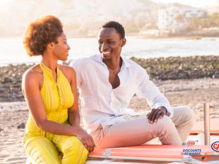 A smiling couple sits on a dock by the water at sunset; she wears a bright yellow outfit, he in a white shirt, enjoying a relaxed moment together.
