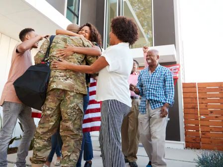 A military member is greeted by a group of cheerful civilians outside a house, sharing hugs and celebration with an American flag.