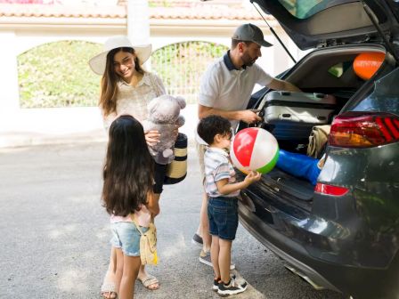 A family loads the car for a trip: kids with toys, a mom holding a stuffed animal, dad near the trunk with beach gear, sunny day.