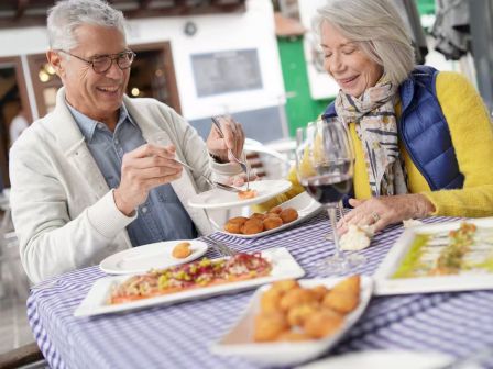 Two seniors enjoying a meal at an outdoor cafe: pizza, bread, and wine on a checkered tablecloth.