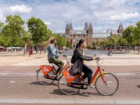 Two people ride a bright orange tandem-style bike on a sunny plaza, with a historic building and trees in the background, enjoying a city ride.