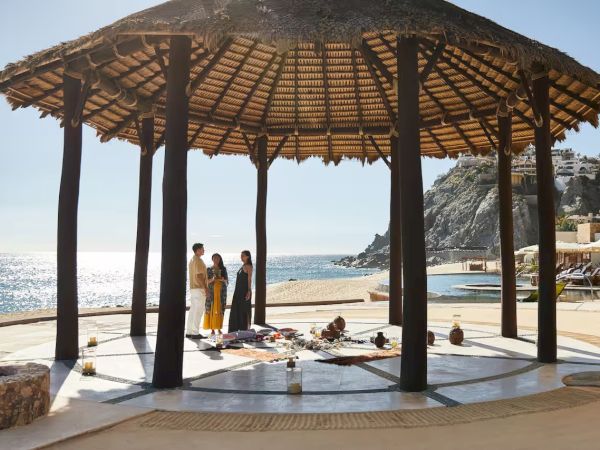 A seaside gazebo with a table set for a meal, two people standing nearby, and an ocean view in the background.