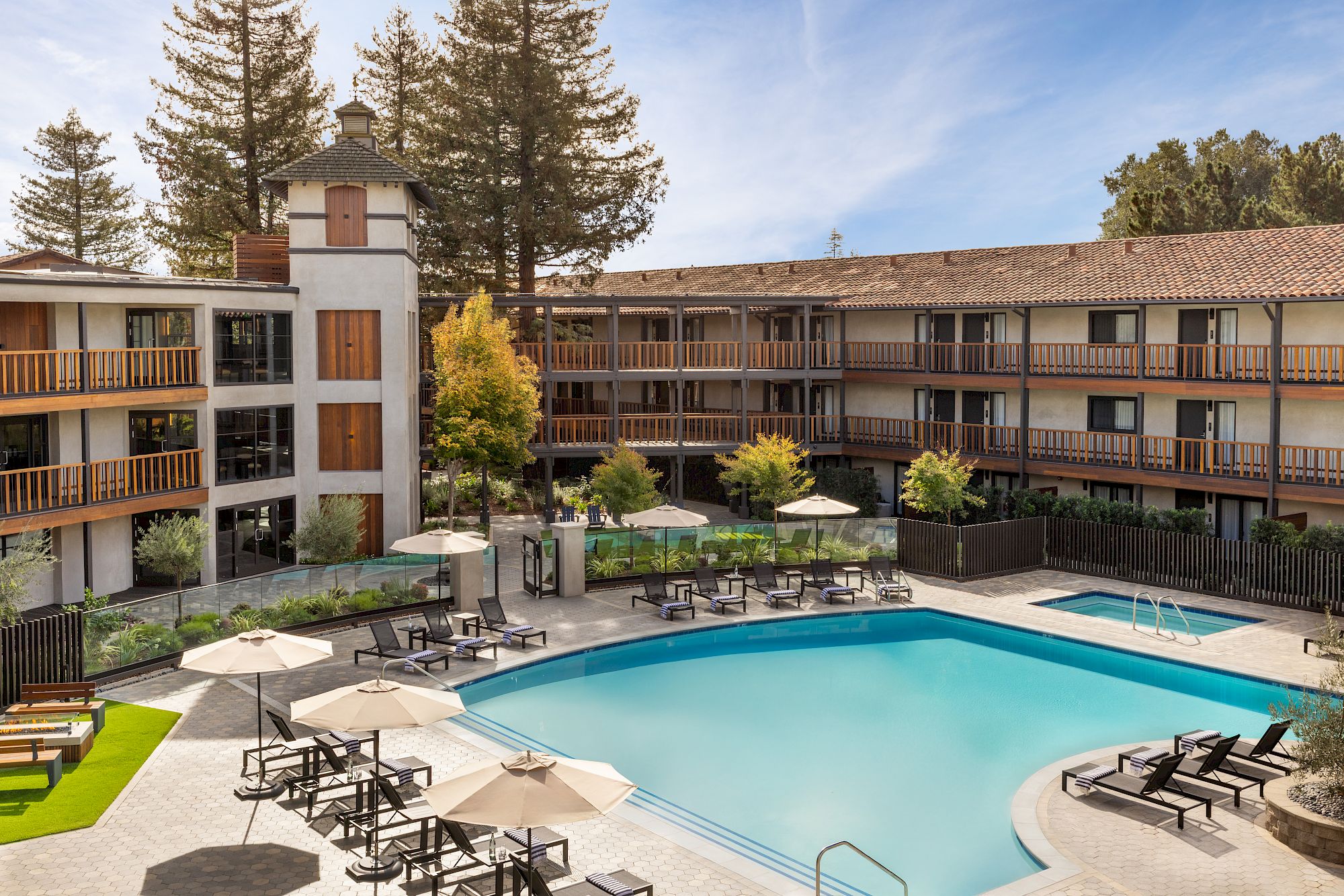 A sunny hotel courtyard with a large rectangular pool, surrounding lounge chairs, umbrellas, and a three-story building with balconies.