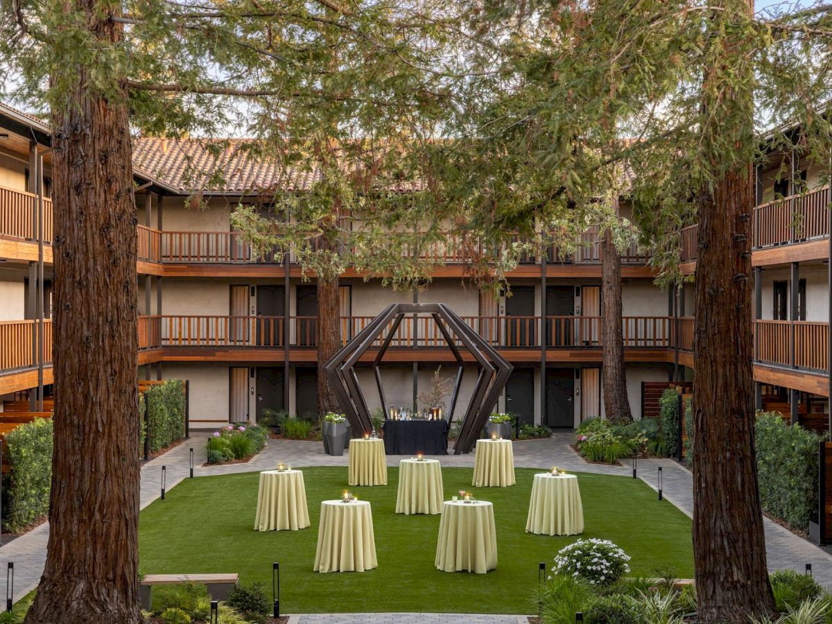 A hotel courtyard with a grassy centerpiece, round tables with tablecloths, and tall trees flanking a central geometric sculpture.