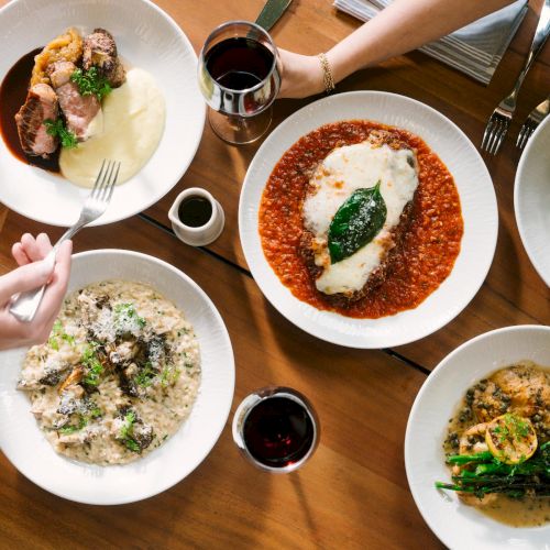 A table of five meals: top-left plate with beef, middle plate with a white sauce, top-right salad, bottom-left creamy risotto, bottom-right greens and grains; glasses nearby.