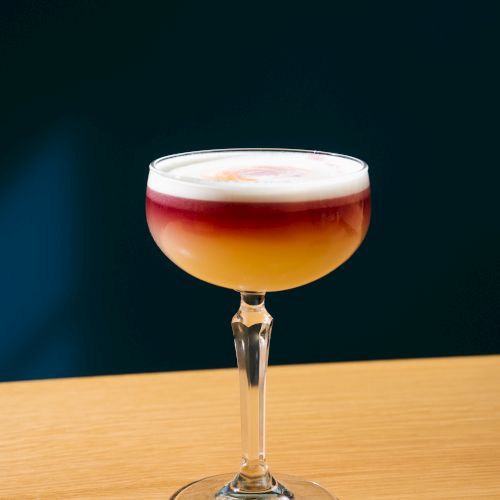 A cocktail in a coupe glass with layered amber drink and a foamy top on a wooden bar, against a dark background.
