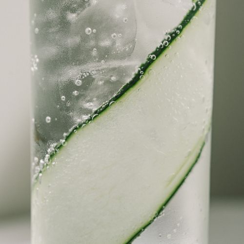 A tall glass of sparkling water with thin lime-green cucumber or lime peels spiraling inside, creating refreshing, clear bubbles.
