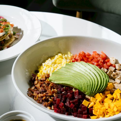 A colorful bowl with rice, avocado slices, corn, black beans, diced beets, peppers, and cheese, plus a small sauce cup beside it.