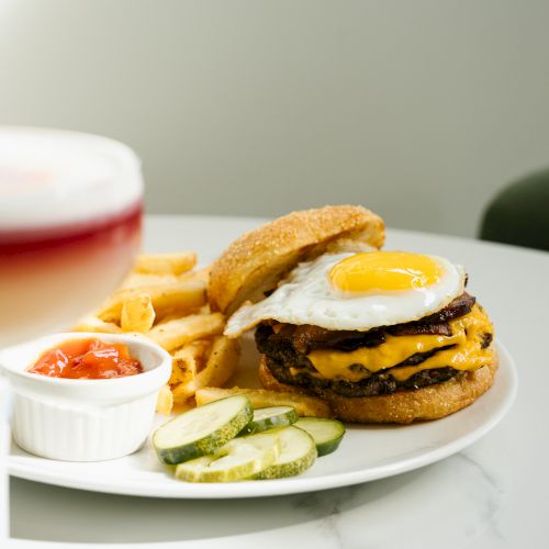 A plate with a bacon cheeseburger topped by a fried egg, beside fries, pickles, and dipping sauces on a white table.