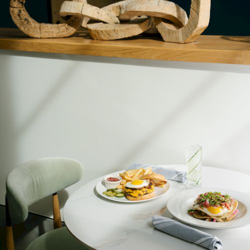 A chic cafe scene with a round table set for two, plates of food and a napkin-wrapped cutlery, plus a large wooden chain sculpture on a shelf above.