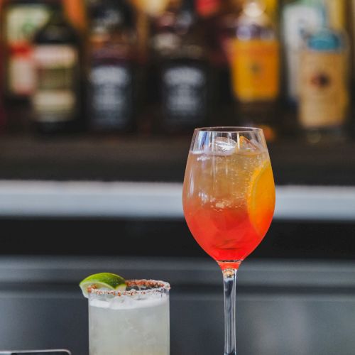 Two cocktails on a bar counter: a light-colored rimmed glass with lime, and a tall orange-to-red layered drink, blurred bottles in the background.