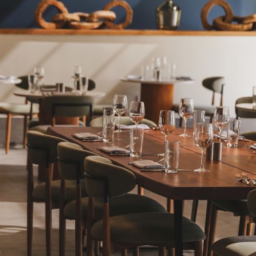 A stylish dining area with wooden tables set for meals, glassware and chairs, calm ambiance, and decorative ropes on a shelf in the background.
