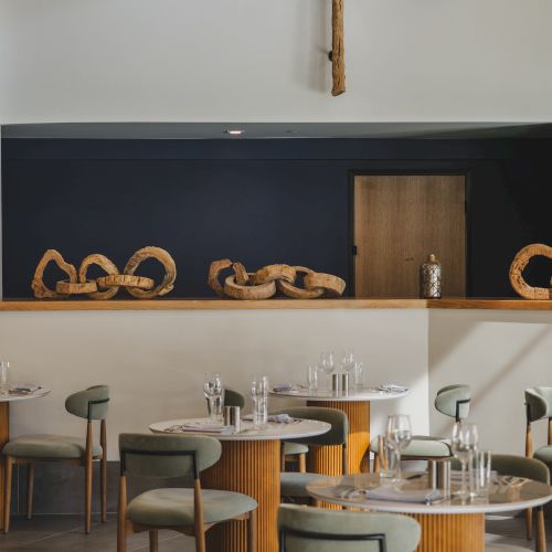 A stylish dining area with wooden barrel tables and gray chairs, decorative twisted branches overhead, and a dark chalkboard wall where looped wooden sculptures rest.