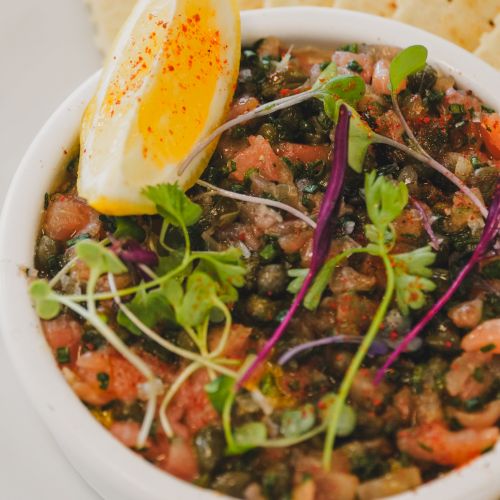 A colorful bowl of pico de gallo-inspired dip with beans and greens, topped with microgreens and a ramekin of lemony oil, served with crackers.