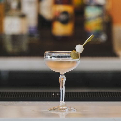 A coupe glass with a pale cocktail, garnished with an olive on a toothpick and a small stick, set against a blurred bar background.