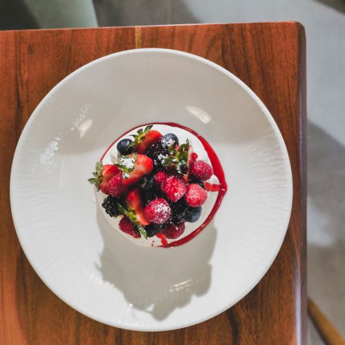A white plate with a berry-topped dessert or smoothie bowl drizzle of red sauce on a wooden table, photographed from above.