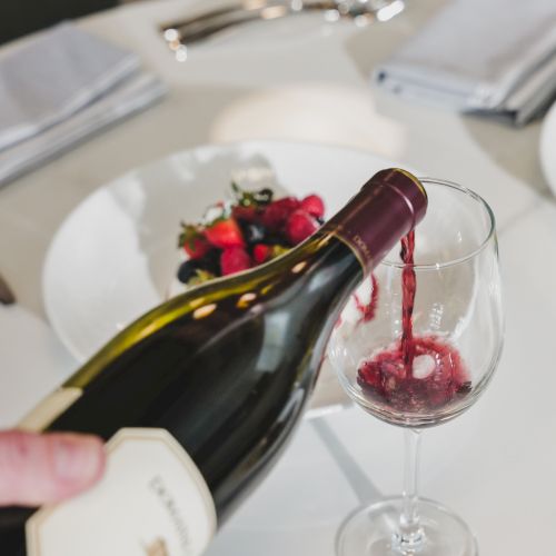 A person pours wine from a bottle into a glass at a table set for dining, with a small bouquet on the plate nearby.