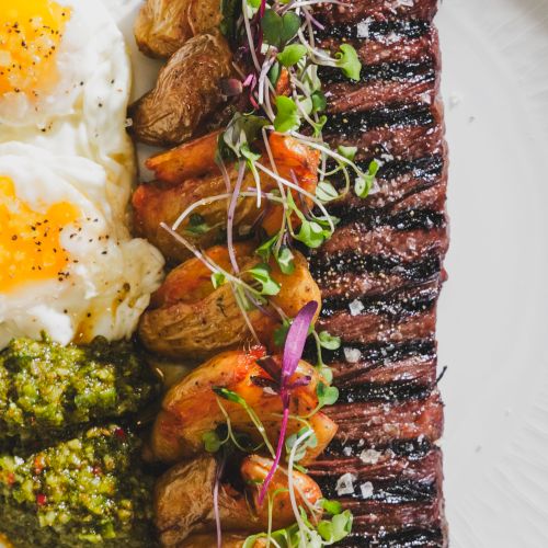 Two sunny-side eggs with pepper on the side, a herb crusted veggie crumble, and a long perfectly grilled steak topped with microgreens and roasted potatoes, all plated elegantly.