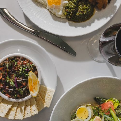 A trio of plates: sunny-side-up eggs with greens and toast, a bowl of avocado/egg salad with crackers, and a fresh veggie pasta salad with boiled eggs, all on a white table.