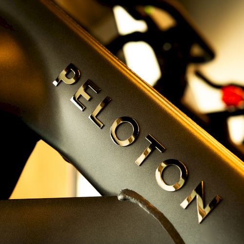 A close-up of a metal frame with the word &ldquo;PELOTON&rdquo; embossed, likely part of a stationary bike, blurred backdrop, warm lighting.