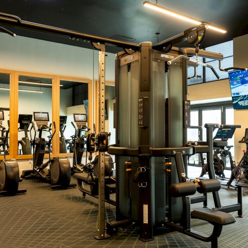 A modern gym with a central multi-gym, cardio machines on the sides, mirrors, and a wall-mounted TV displaying a workout.