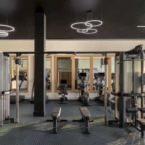 A modern gym with weight machines and cardio equipment, plus glowing ring pendant lights overhead.