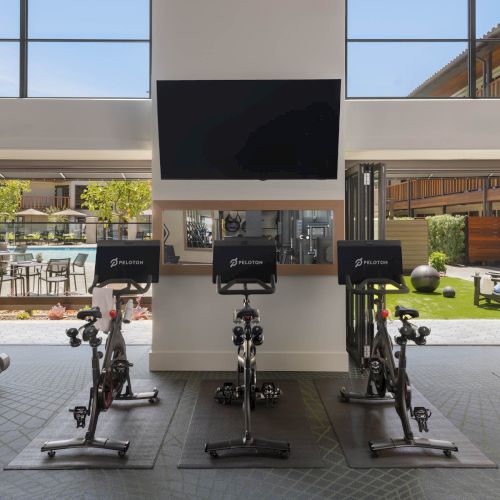 A modern courtyard gym with several Peloton bikes, big windows, and an outdoor lounge by a lawn and pool area.