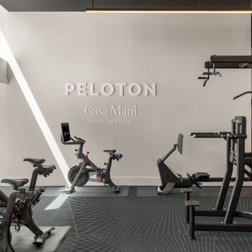 A modern hotel gym with Peloton bikes, a fitness machine, and the wall text &ldquo;PELoton Casa Mani Resort Napa Valley,&rdquo; neatly lit by ceiling lights.