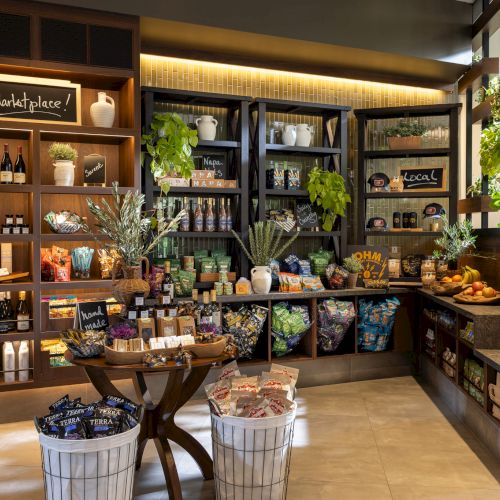 A warm-lit grocery/market boutique with shelves of wine, snacks, jars, and plants, plus a chalkboard reading &ldquo;The Marketplace!&rdquo;.