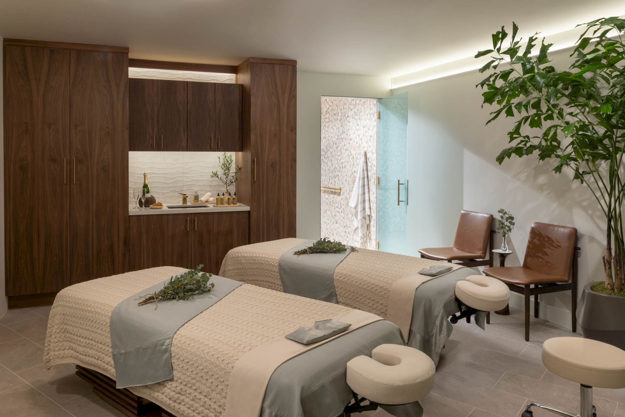 A modern spa room with treatment beds, wooden cabinetry, massage chairs, and a leafy plant near the window.