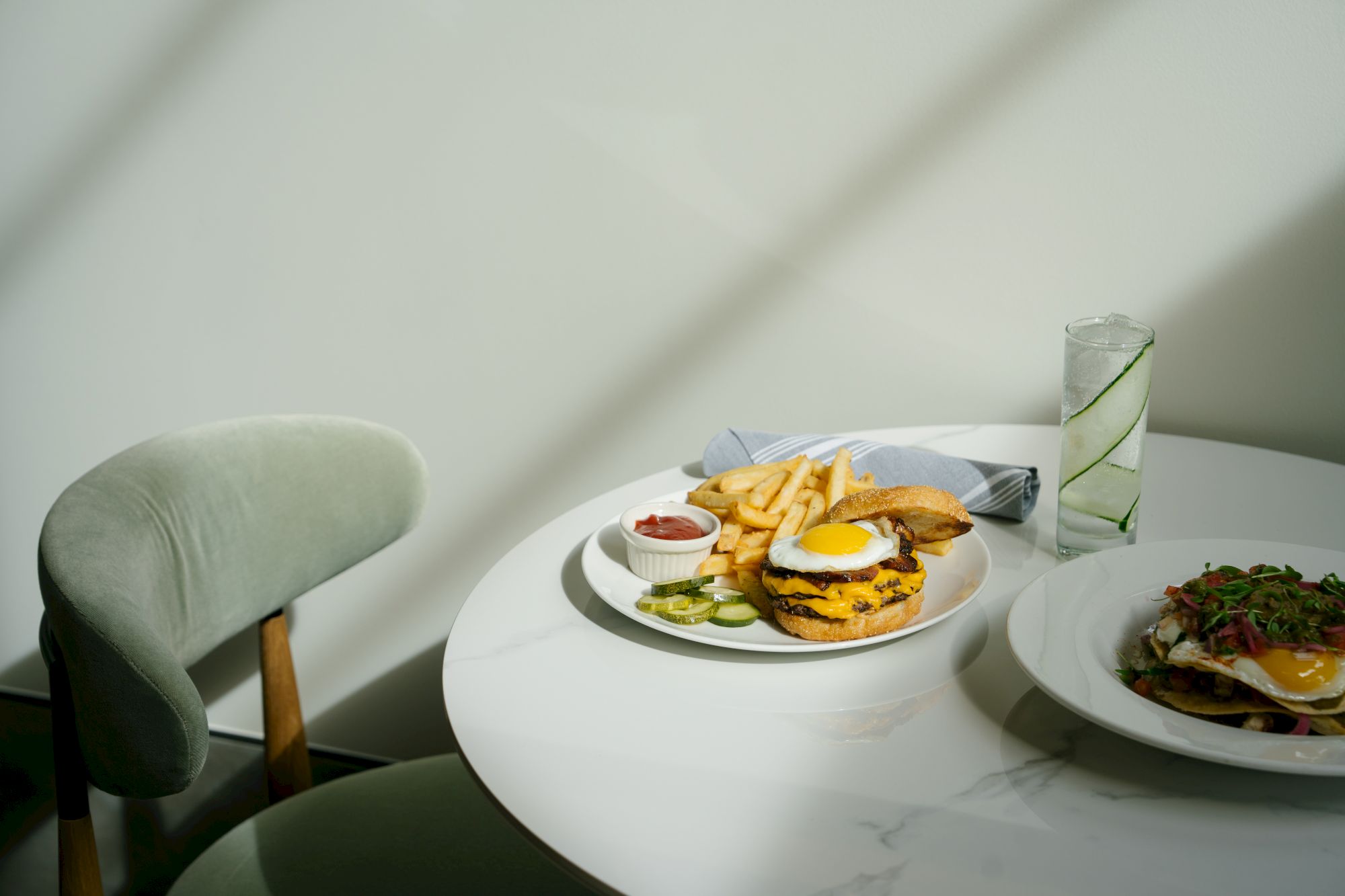 A dining setup with two plates: one with a burger, fries, and dipping sauces; the other with a steak and greens, on a white round table next to a chair.