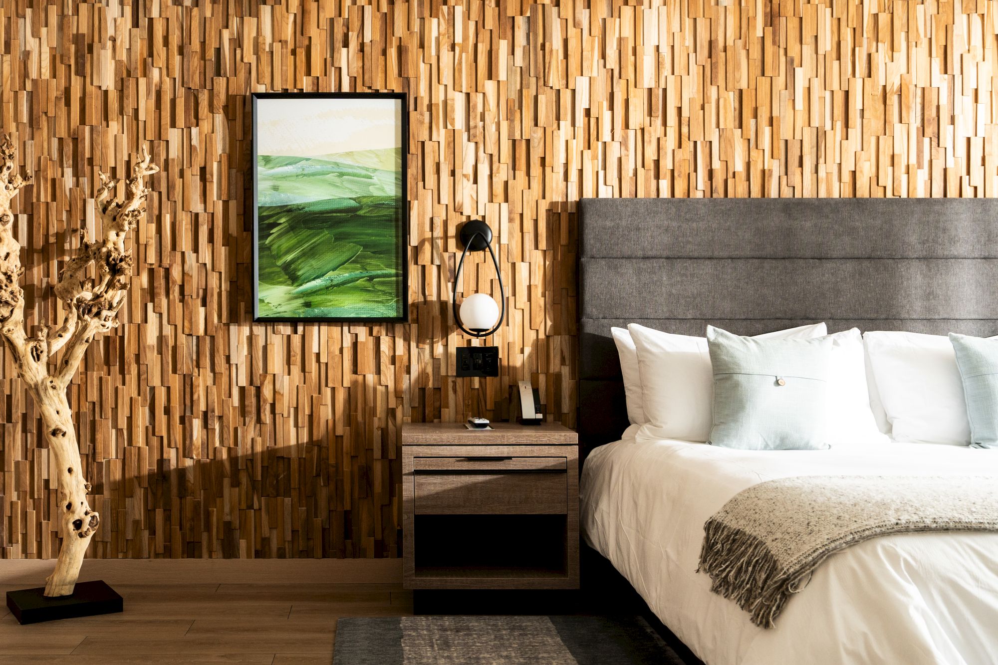 A modern bedroom with a wood-textured feature wall, a gray upholstered headboard, a nightstand with lamp, framed green landscape art, and a small tree sculpture.