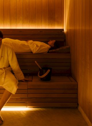 A person wearing a yellow shirt sits at a wooden bench or steps, holding a dark object against their leg with hands resting nearby.