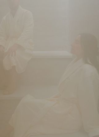 Two people in light robes sit and stand in a softly lit spa or bathhouse, robes draped, misty peaceful ambiance.