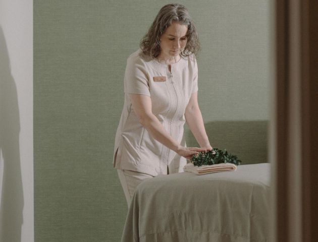 A woman sits on a bed in a vintage bedroom, wearing a light pink robe; soft light, muted green walls, and a calm, nostalgic mood.
