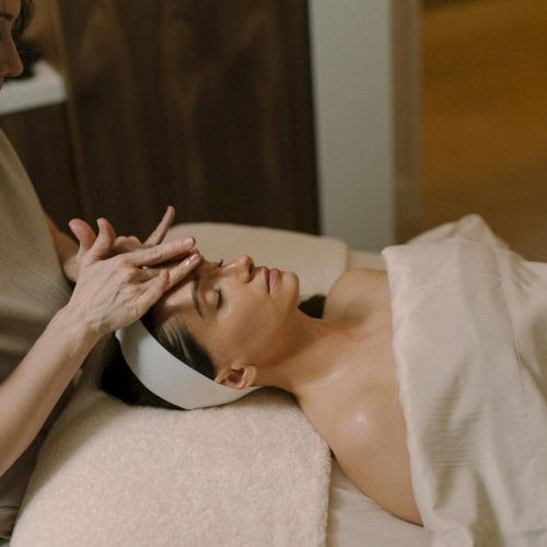 A person is receiving a facial massage with hands resting near the forehead while lying on a treatment bed.