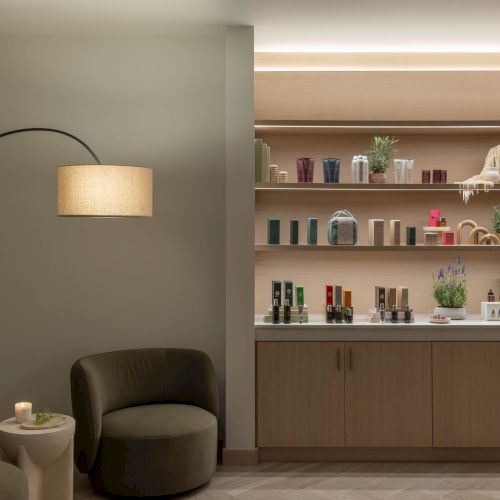A modern spa lounge with a floor lamp and open shelves stocked with skincare bottles and small plants.